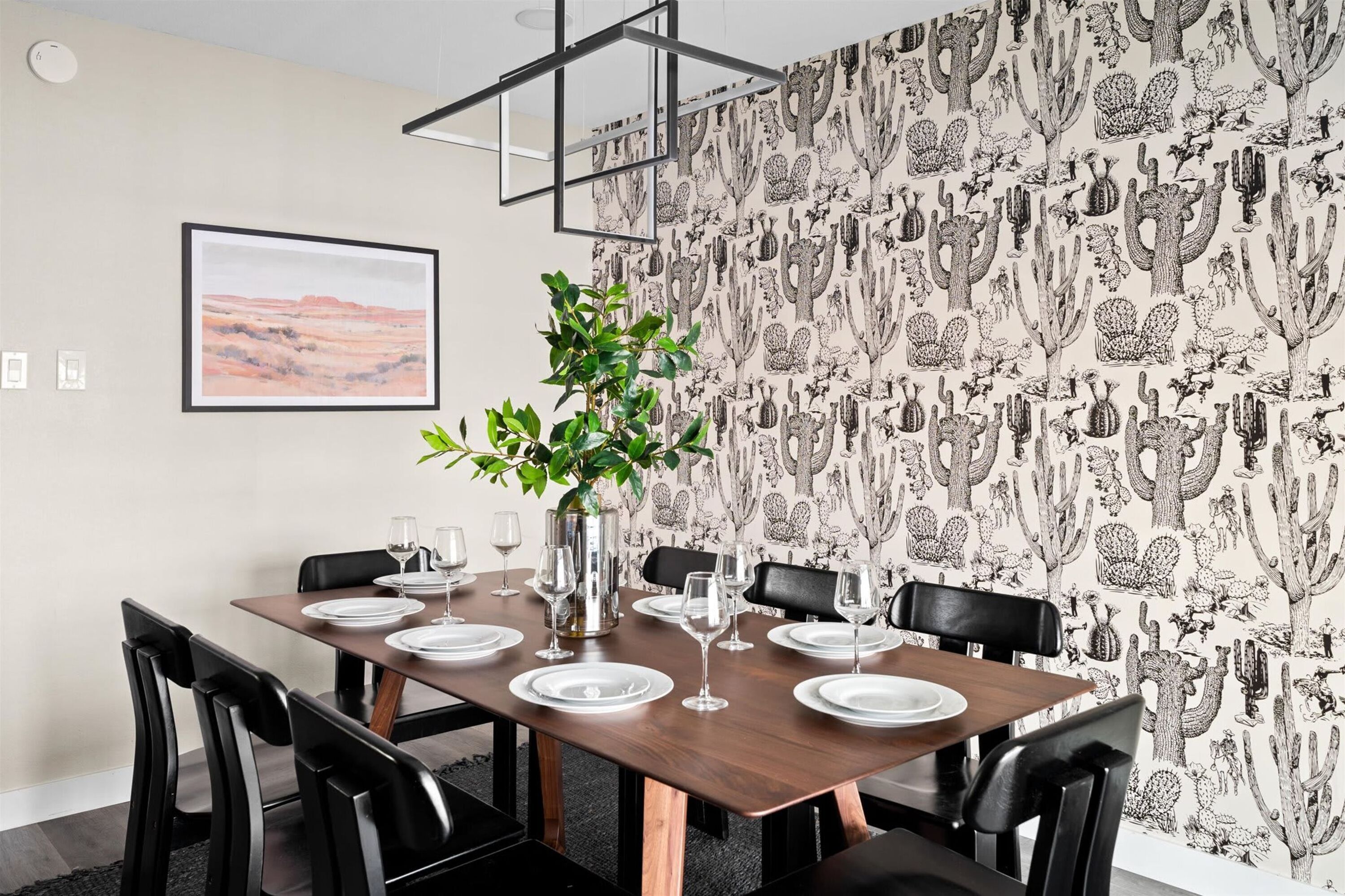 Un coin repas avec une table en bois dressée pour quatre personnes, entourée de chaises noires, d’une plante en pot et d’un tableau encadré accroché au mur.