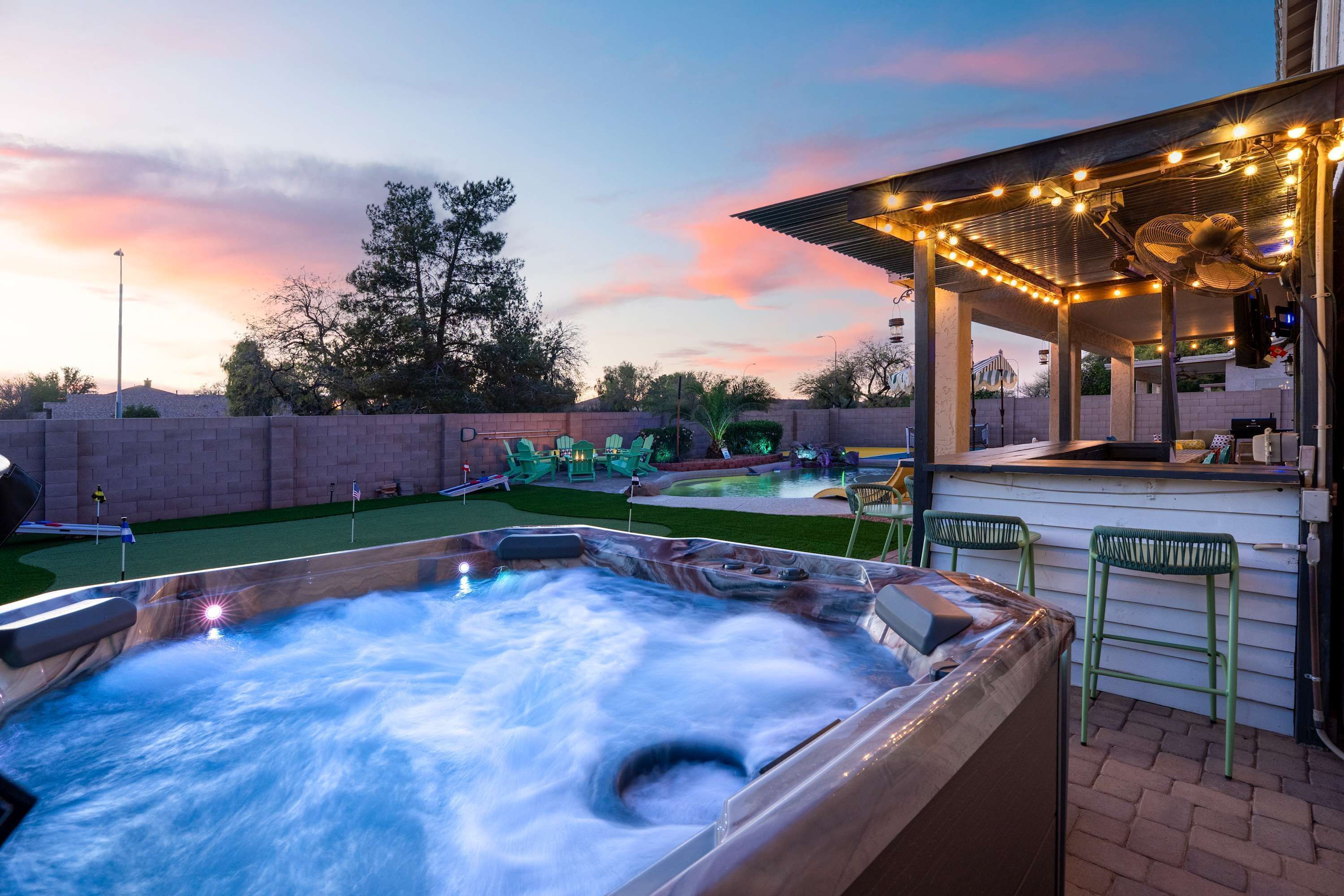 A hot tub with a waterfall feature, a covered outdoor seating area with string lights, and a pool with a water slide.