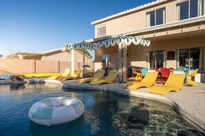 A swimming pool with lounge chairs and a floating ring, surrounded by a building with large windows and a covered patio area. - Bloomin Dunes - Pool Pickleball Tiki Bar (Chandler)