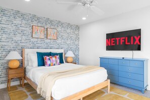 A bedroom with a bed, two nightstands, a blue dresser, a ceiling fan, and a wall-mounted TV displaying 'NETFLIX'. - Bloomin Dunes - Pool Pickleball Tiki Bar (Chandler)