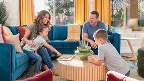 Eine vierköpfige Familie in einem Wohnzimmer mit einem blauen Sofa, einem Holztisch und Blick ins Freie durch große Fenster.