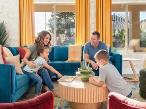 A family of four in a living room with a blue sofa, a wooden coffee table, and a view of the outdoors through large windows. - Bloomin Dunes - Pool Pickleball Tiki Bar (Chandler)