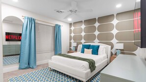 Ein modernes Schlafzimmer mit Bett, Vorhängen, Deckenventilator, Wand-Fernseher und einem gemusterten Teppich.