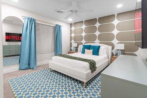 A modern bedroom with a bed, curtains, a ceiling fan, a wall-mounted TV, and a patterned rug. - Bloomin Dunes - Pool Pickleball Tiki Bar (Chandler)