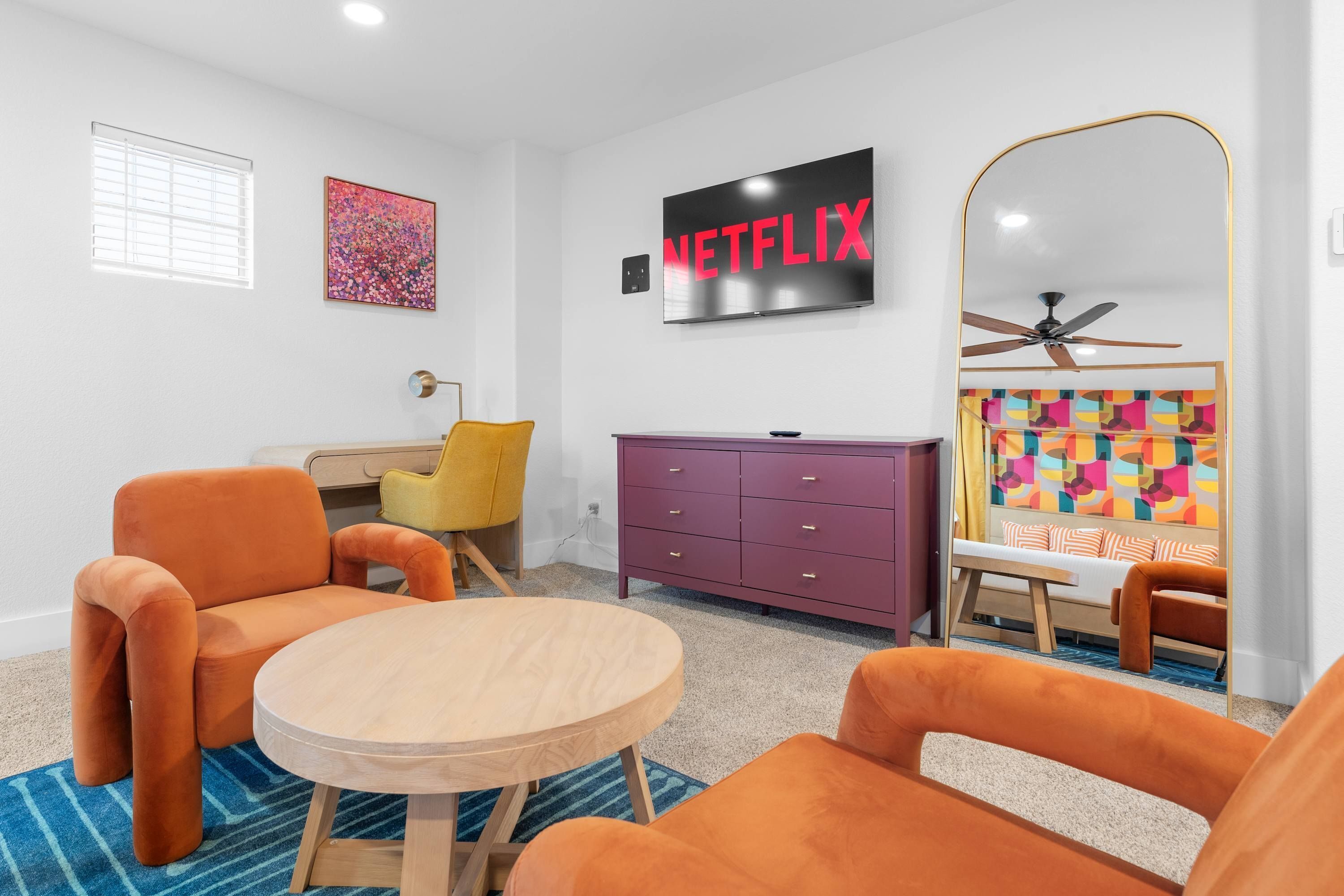 A modern living room with a round wooden table, orange armchairs, a purple dresser, a ceiling fan, and a wall-mounted TV displaying 'NETFLIX'.