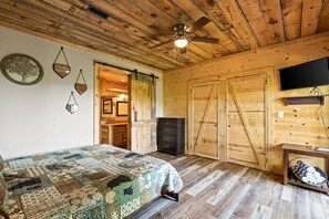 Un dormitorio con una cama, ventilador de techo, televisión y paredes de madera.