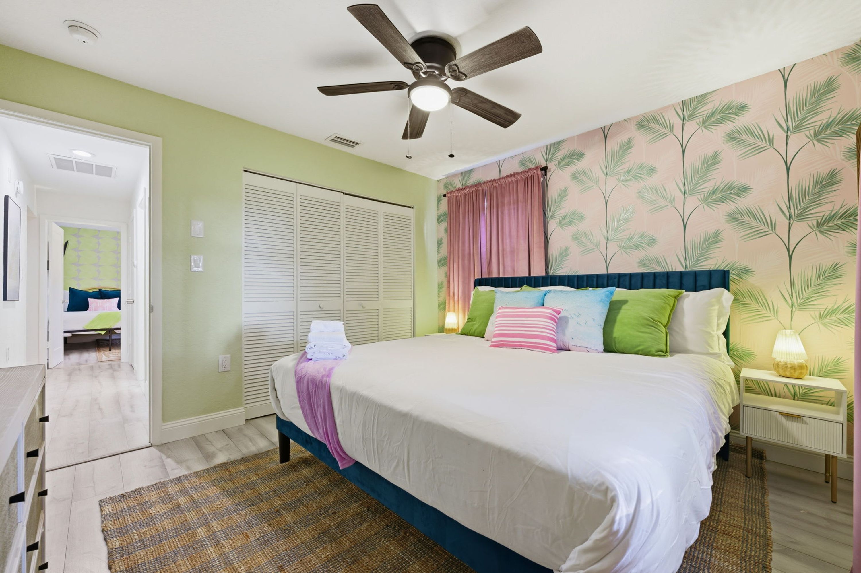Une chambre à coucher avec un lit, un ventilateur de plafond, une table de chevet, une lampe et une fenêtre avec des rideaux.