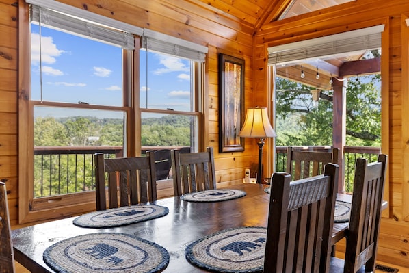 A wooden interior with a dining table set for six, surrounded by chairs, large windows offering a view of the outdoors, and a lamp on the table. - Mountain Views Hot Tub Pool Table Firepit Games (Epworth)
