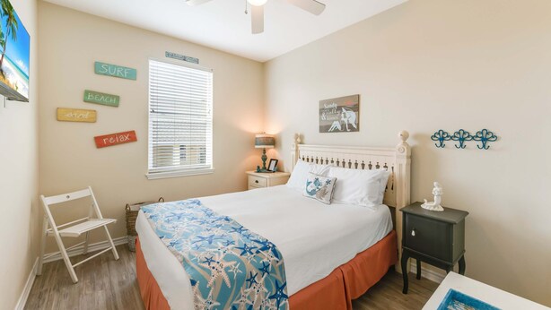 Une chambre à coucher comprenant un lit, une table de chevet, une chaise, un ventilateur de plafond et des éléments de décoration murale, notamment un panneau sur le thème de la plage et une illustration de sirène.