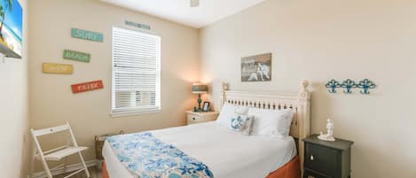 Une chambre à coucher comprenant un lit, une table de chevet, une chaise, un ventilateur de plafond et des éléments de décoration murale, notamment un panneau sur le thème de la plage et une illustration de sirène.