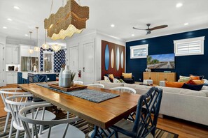 A modern living room with a dining area, featuring a wooden dining table, white chairs, a blue sofa, and a ceiling fan. - Hilton Head 6BR Sleeps 18 Pool Hot Tub Firepit (Hilton Head Island)