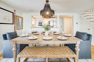 Comedor con mesa de madera dispuesta para seis personas, flanqueada por dos sofás grises, un banco tejido y una araña.