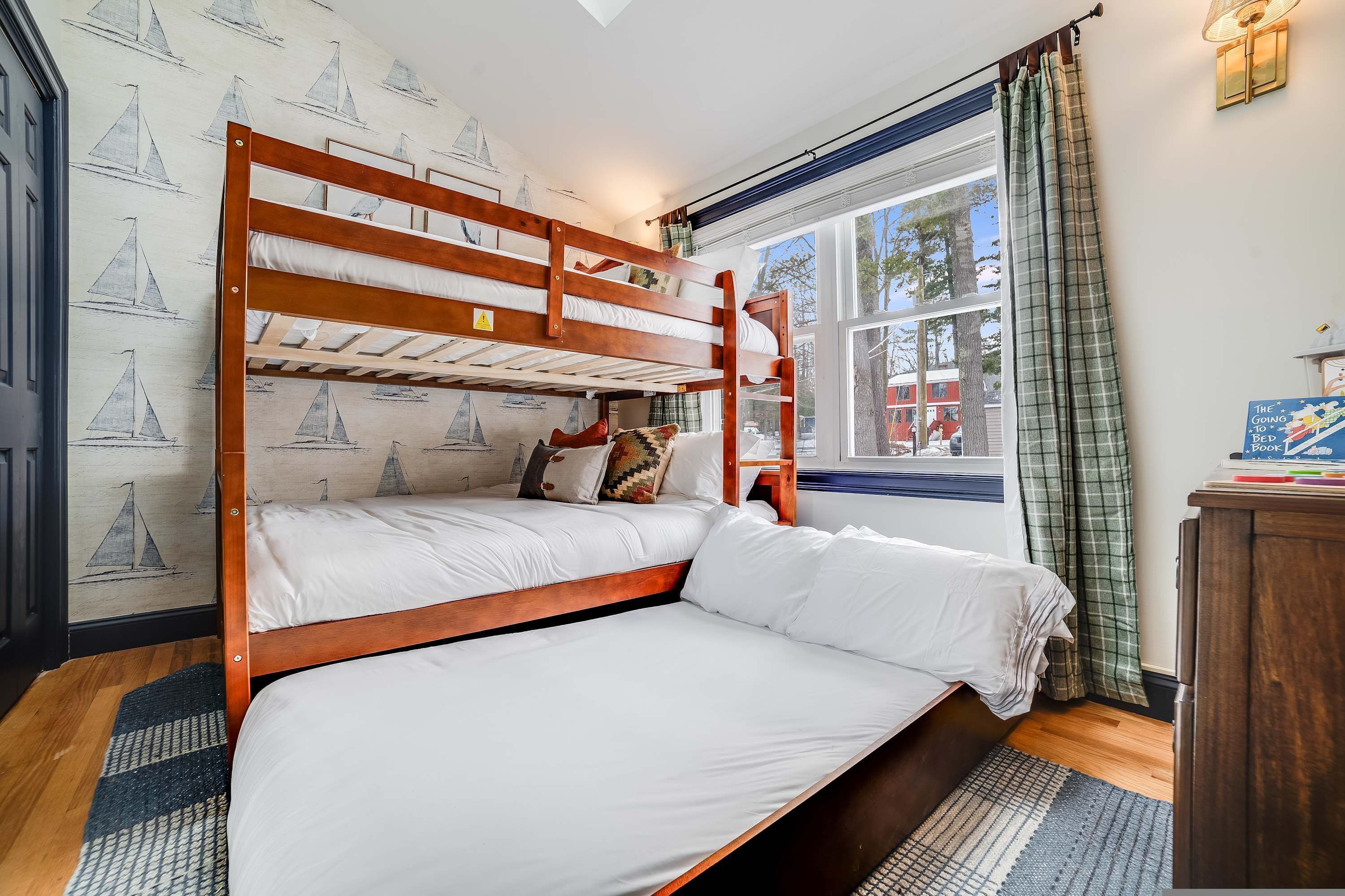A bunk bed room with a window, a wall with sailboat wallpaper, and a bedside table with books.