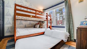 A bunk bed room with a window, a wall with sailboat wallpaper, and a bedside table with books.