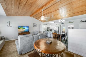 Een moderne woonkamer met een houten plafond, een televisie, grijze banken, een houten eettafel en een keuken op de achtergrond.