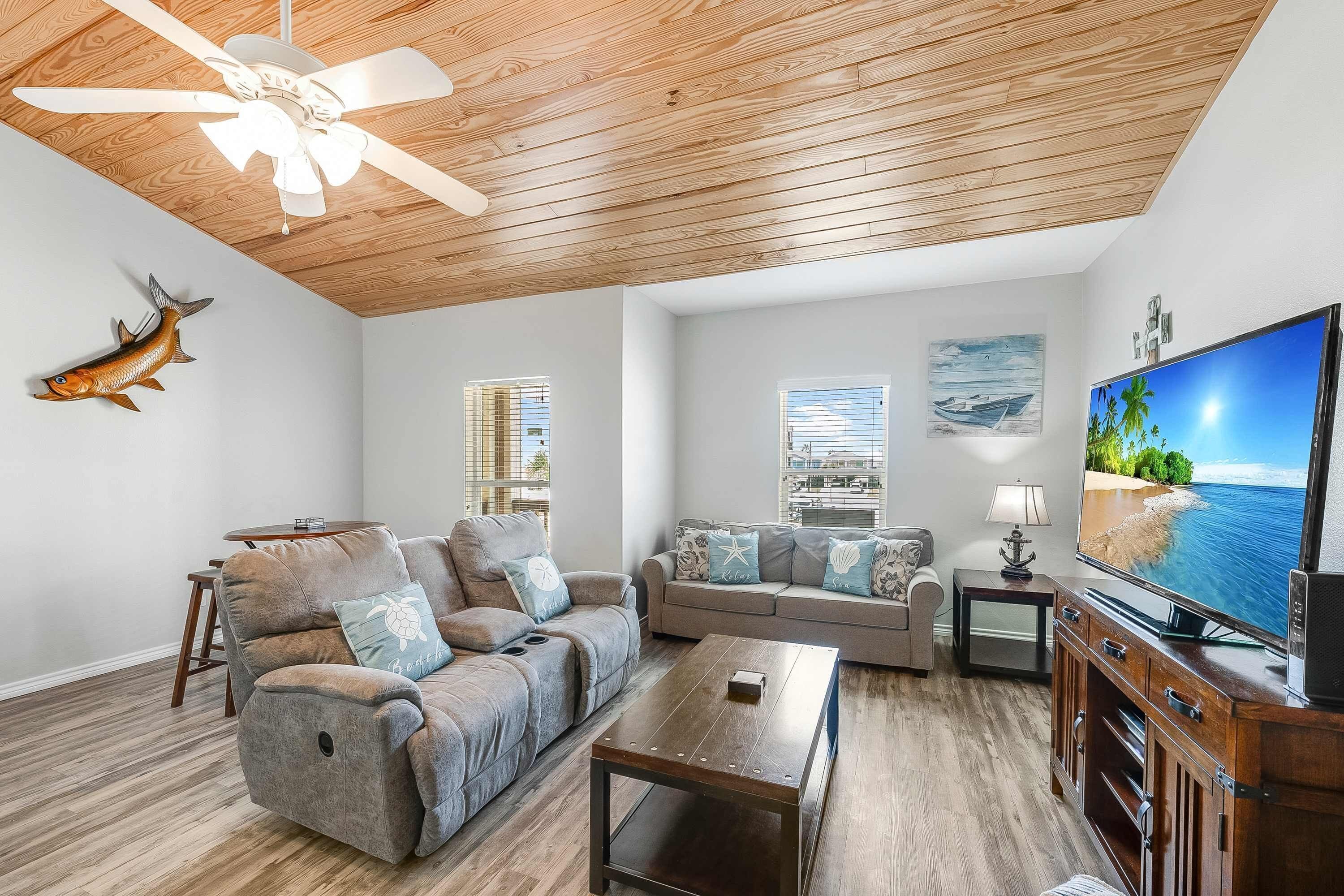 A living room with a ceiling fan, a fish decoration, a sofa set, a television, and a view of the sea through the windows.