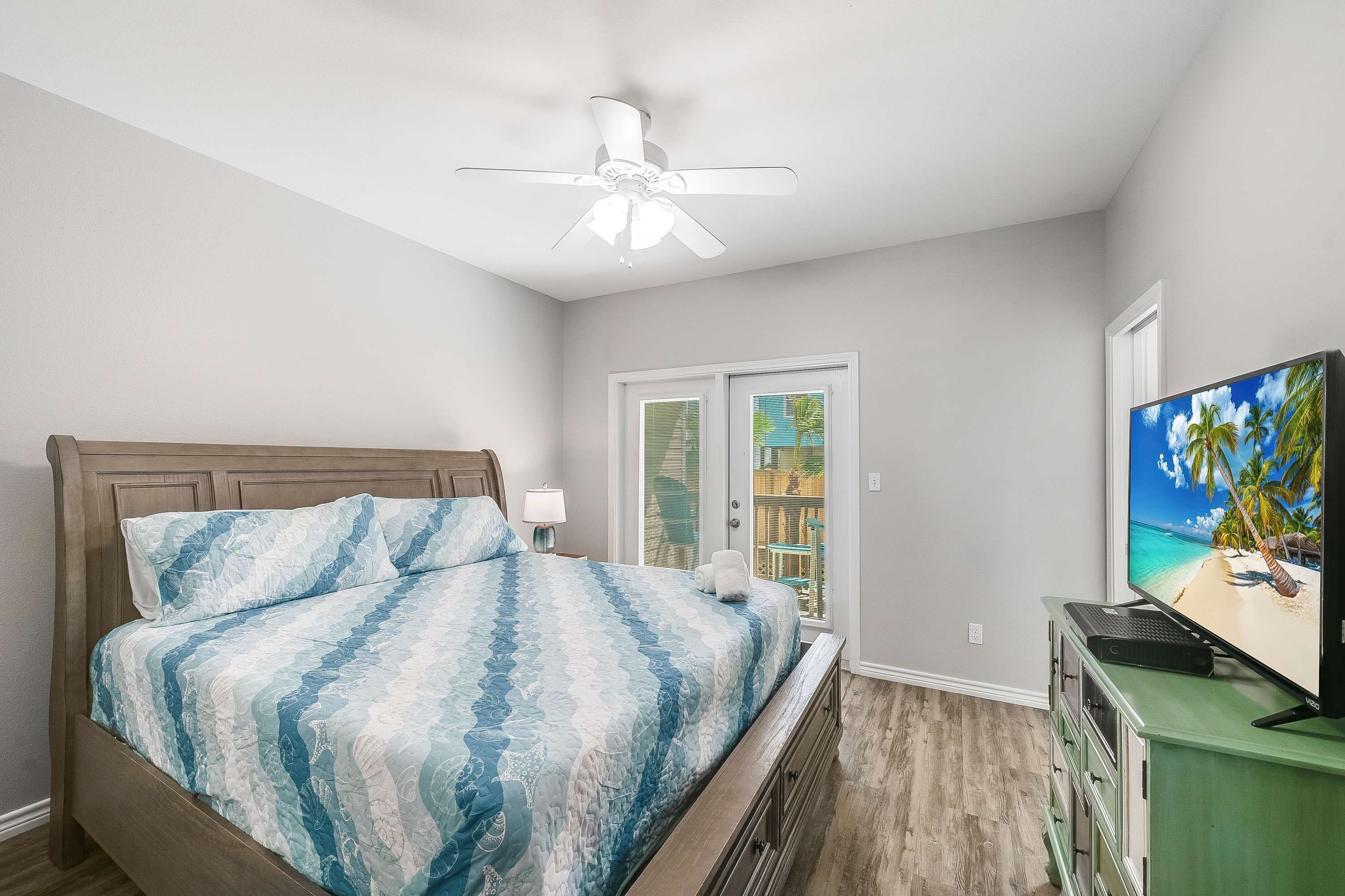 A bedroom with a bed, a ceiling fan, a television, and a view of the outdoors through a door.