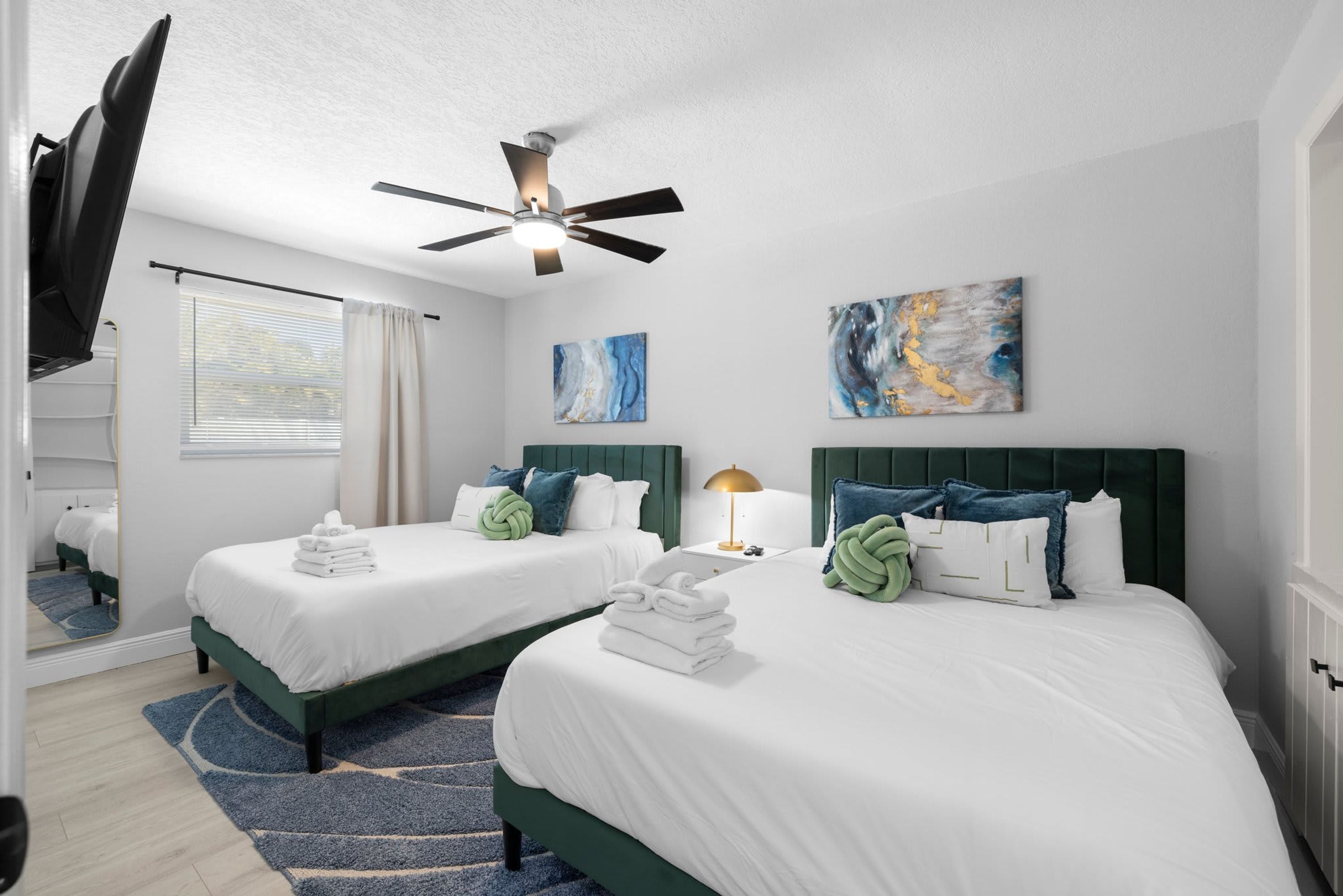 A hotel room with two beds, a ceiling fan, artwork on the walls, and a window with blinds.