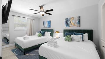 A hotel room with two beds, a ceiling fan, artwork on the walls, and a window with blinds.