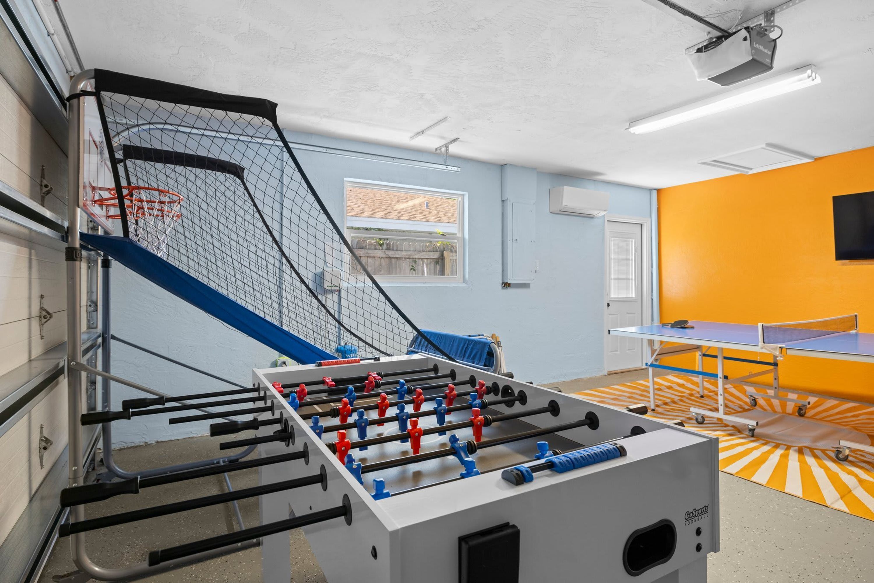 A room with a foosball table, a ping pong table, and a basketball hoop.