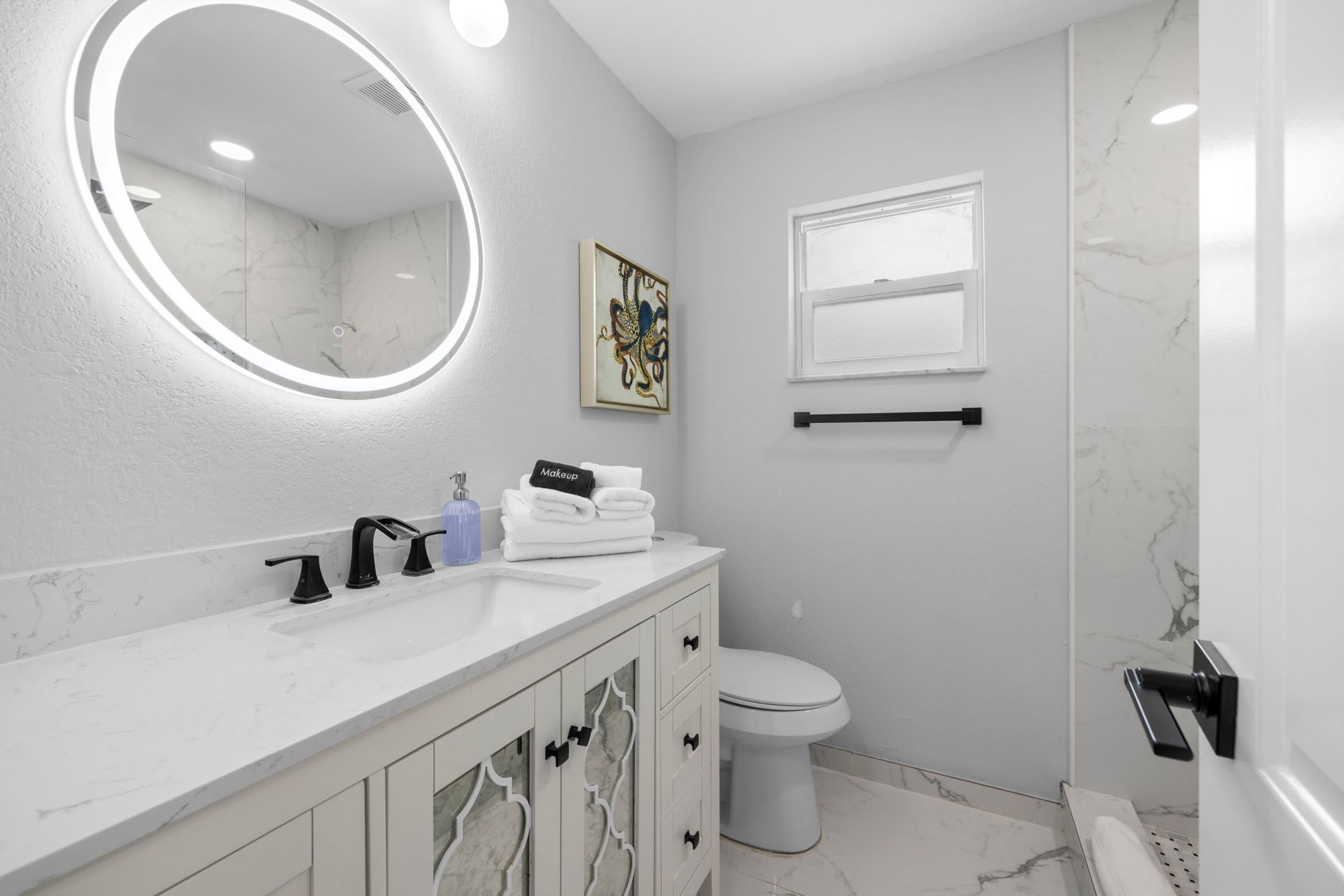 Un baño moderno con un tocador blanco, un espejo redondo, un inodoro y una zona de ducha.