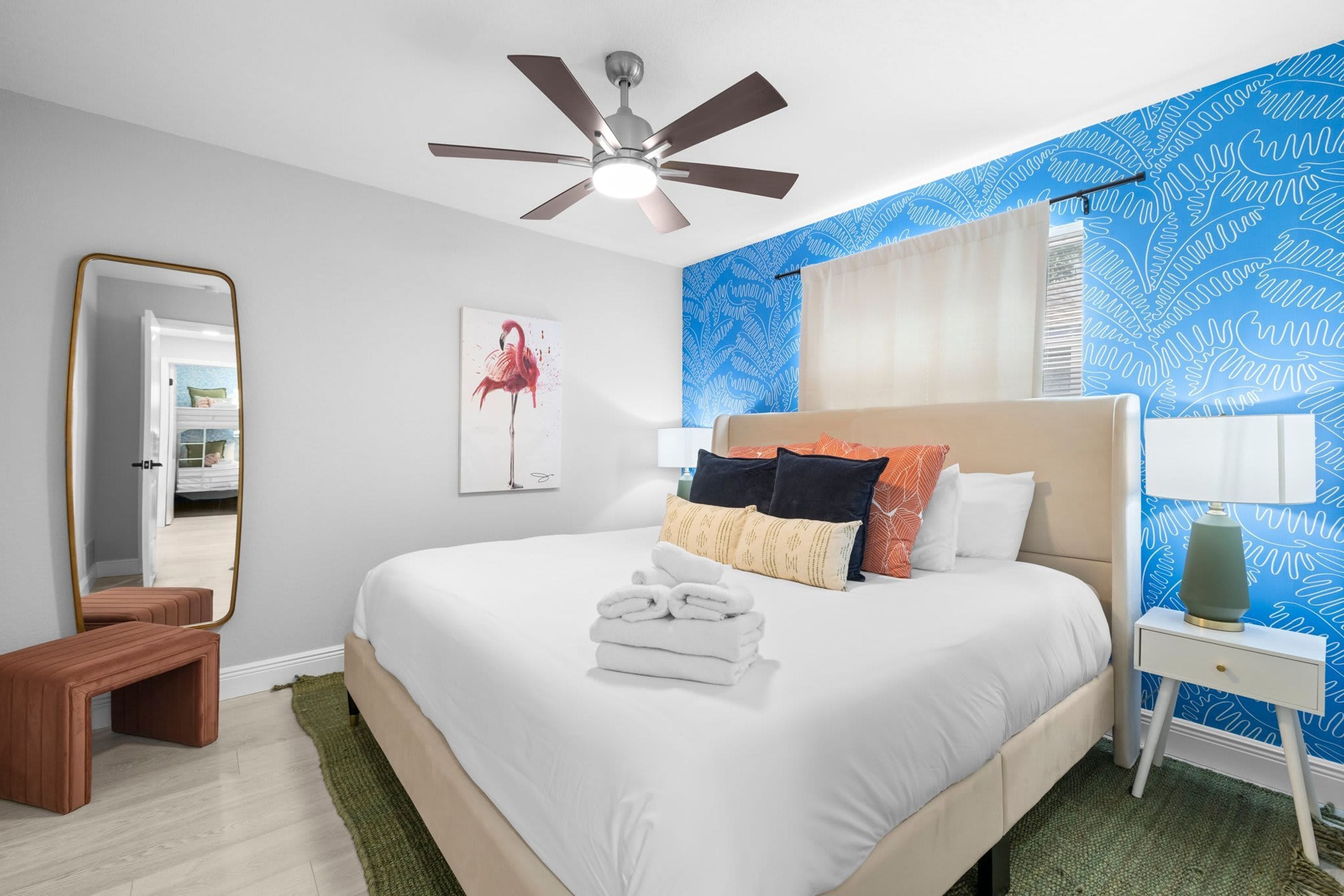 A hotel room with a bed, a ceiling fan, a nightstand with a lamp, a mirror, and a blue patterned wall.