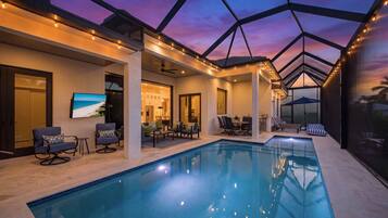 A modern indoor swimming pool with a glass ceiling, surrounded by lounge areas and a kitchen.