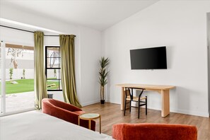 A modern hotel room with a bed, a desk, a chair, a television, and a view of a garden. - The Lazy Palm Private Lazy River Pickleball (Glendale)