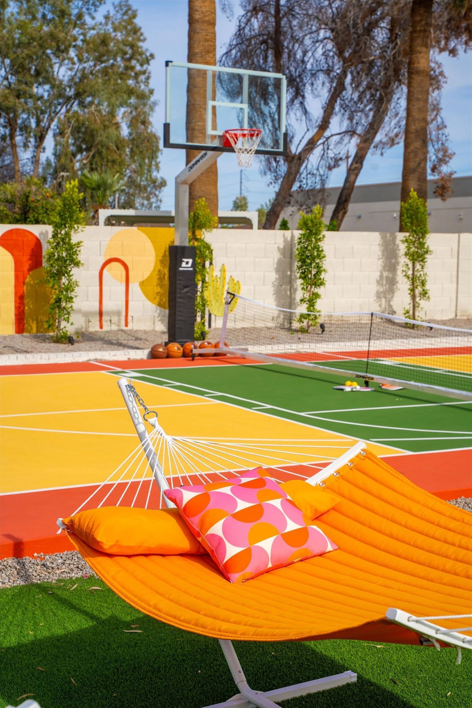 Ein Basketballplatz mit einer Hängematte und Kissen, umgeben von Bäumen und einer Wand mit farbenfrohen Mustern.