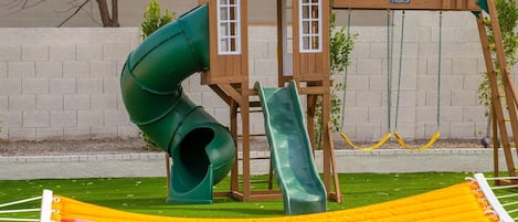 A playground with a slide and swings, a hammock, and a green playhouse.