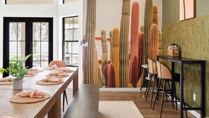 A modern dining area with a long table set for a meal, a cactus wall mural, and a view of the outdoors through large windows. - The Lazy Palm Private Lazy River Pickleball (Glendale)