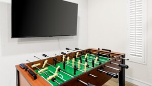 A room with a foosball table, a television mounted on the wall, and a window with blinds. - The Lazy Palm Private Lazy River Pickleball (Glendale)