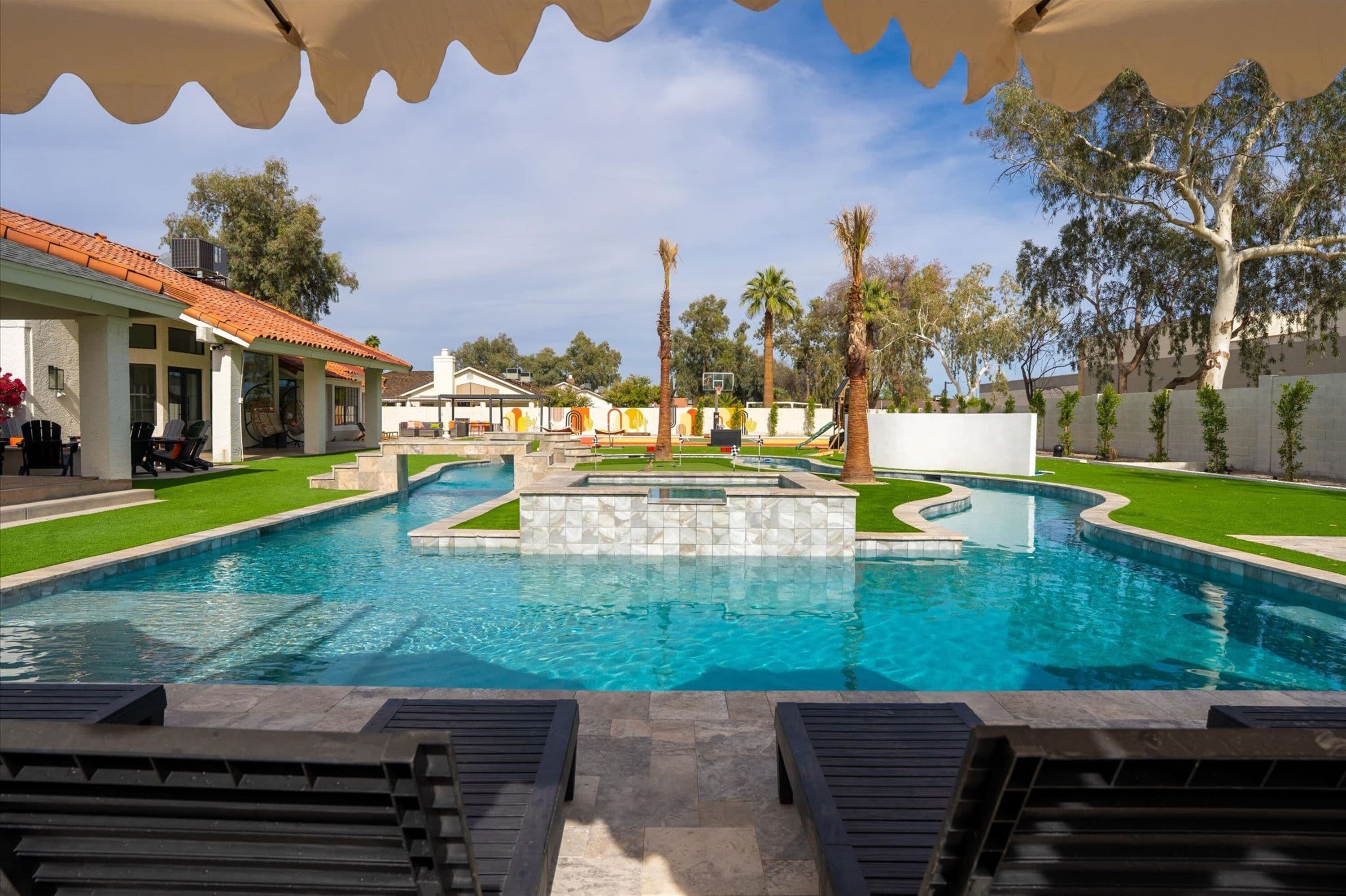 A swimming pool with a tiled deck, lounge chairs, and a covered area with a wavy roof.