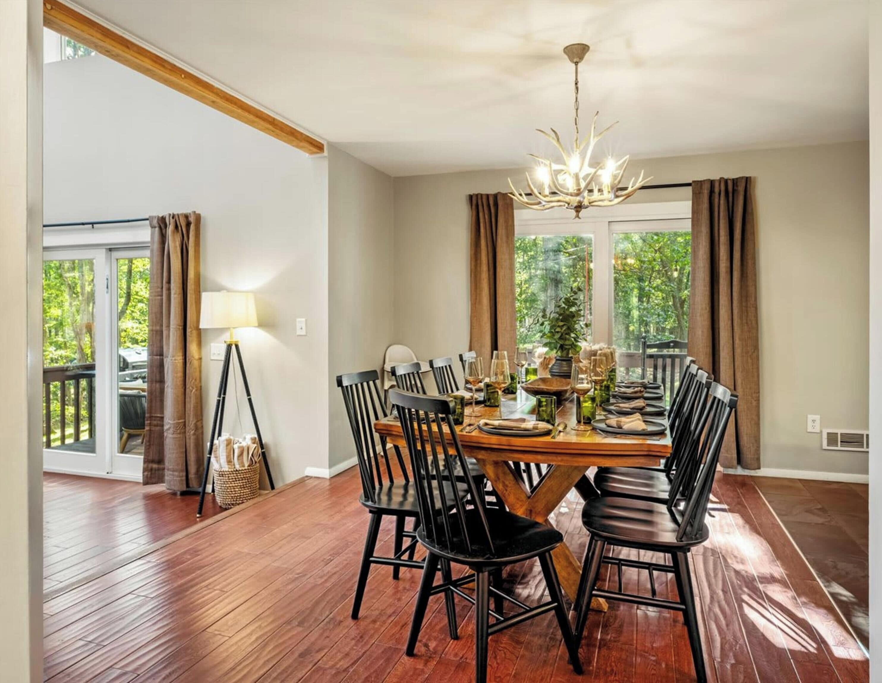 A dining room with a wooden table set for six, surrounded by black chairs, a chandelier, and large windows with curtains.