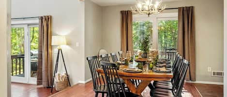 Comedor con una mesa de madera dispuesta para seis personas, rodeada de sillas negras, un candelabro y ventanas grandes con cortinas.