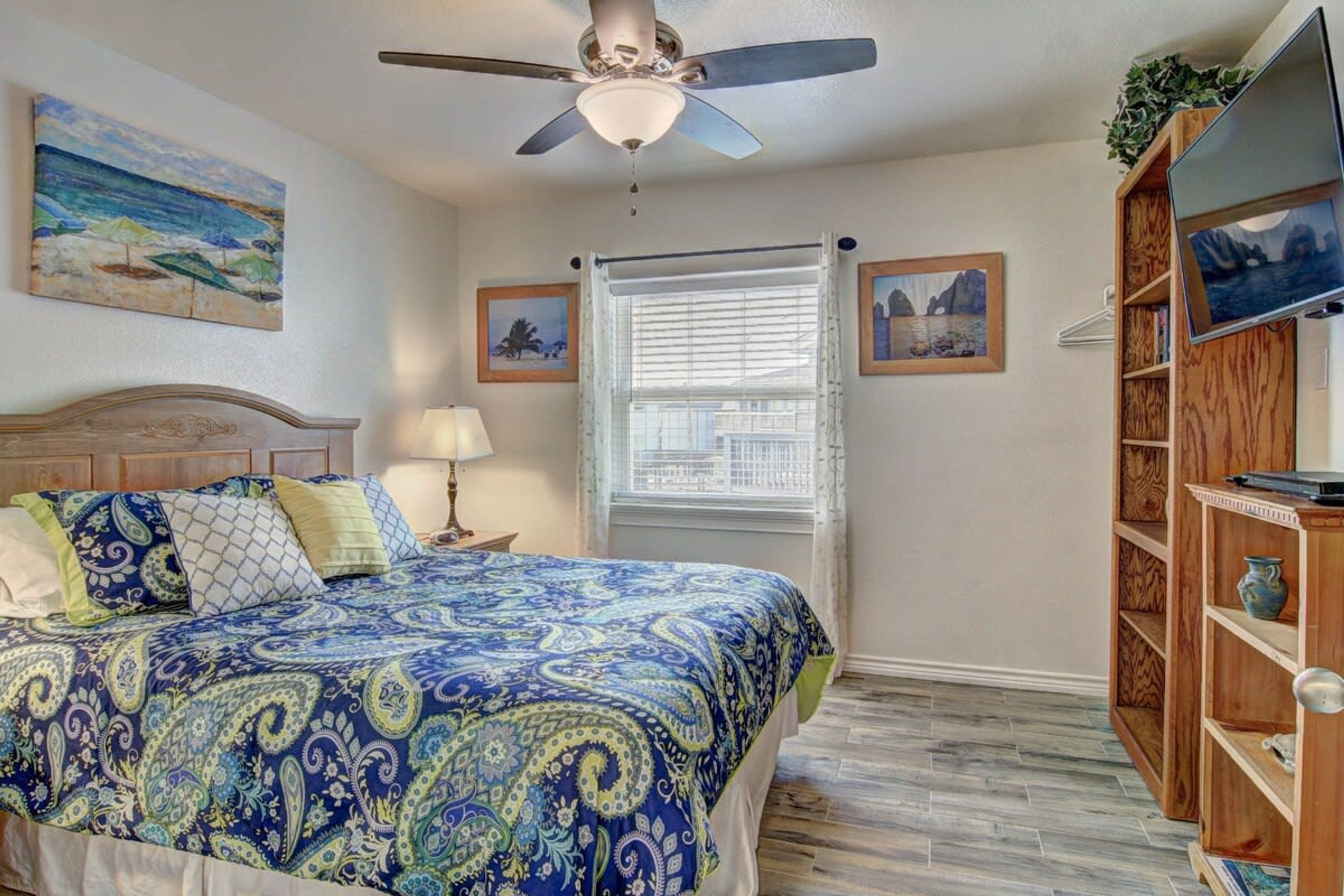 A bedroom with a bed, a ceiling fan, a window with blinds, and a television on a wooden stand.