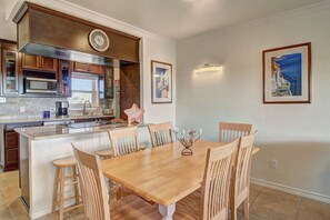 A dining area with a wooden table and chairs, a kitchen with wooden cabinets and a microwave, and two framed pictures on the wall. - Buena Vista- Ocean View Large Pool Boardwalk (Port Aransas)