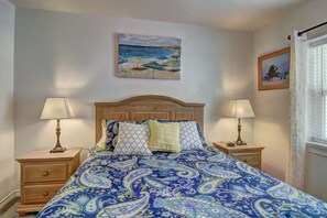 A bedroom with a bed, two bedside tables, a nightstand, a window with curtains, and two wall-mounted paintings. - Buena Vista- Ocean View Large Pool Boardwalk (Port Aransas)