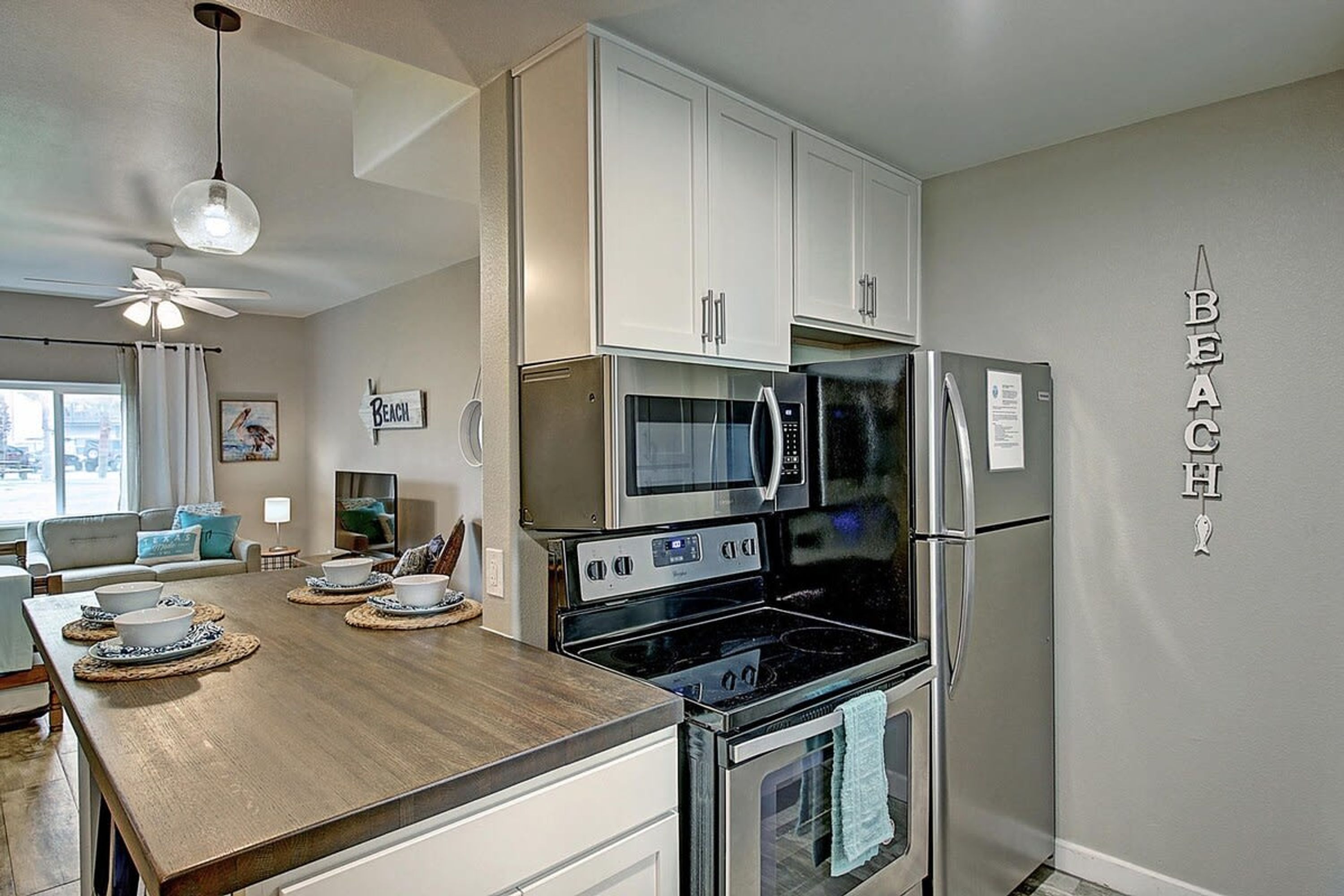 A modern kitchen with white cabinets, a microwave, a refrigerator, and an oven. There is a wooden dining table with bowls and a coffee maker.