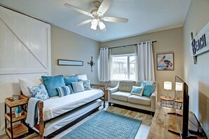 A bedroom with a bed, a sofa, a ceiling fan, a window with curtains, and a wall-mounted sign. - Isle Getaway Boardwalk to Beach Community Pool (Port Aransas)