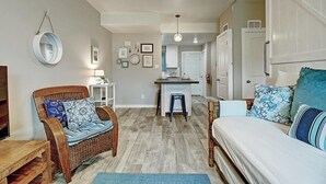 Um quarto aconchegante com uma cama, uma cadeira de vime, uma mesinha com abajur, e uma cozinha compacta ao fundo.
