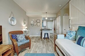A cozy room with a bed, a wicker chair, a small table with a lamp, and a kitchenette in the background. - Isle Getaway Boardwalk to Beach Community Pool (Port Aransas)