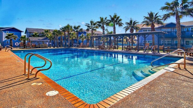 Uma piscina com deck de madeira, palmeiras e um edifício com fachada azul.