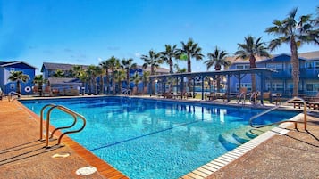 A swimming pool with a wooden deck, palm trees, and a building with a blue facade.