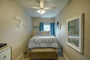 A bedroom with a bed, a ceiling fan, a dresser, a window with curtains, and a mirror. - Isle Getaway Boardwalk to Beach Community Pool (Port Aransas)