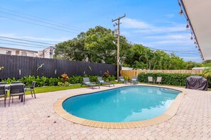 A swimming pool with lounge chairs and a table, surrounded by a wooden fence and greenery. - 2 Miles to Beach Pool Cabana Pool Table (Deerfield Beach)