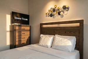 Um quarto com cama, cômoda, televisão e luminária na parede.