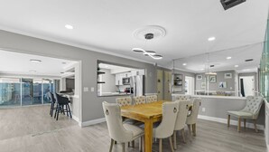 A modern dining area with a wooden table and upholstered chairs, a kitchen in the background, and a glass partition separating the dining area from another space. - Sleeps 8 Tiki Bar Pool Hot Tub Fire Pit (Tampa)
