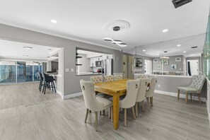 A modern dining area with a wooden table and upholstered chairs, a kitchen in the background, and a glass partition separating the dining area from another space. - Sleeps 8 Tiki Bar Pool Hot Tub Fire Pit (Tampa)