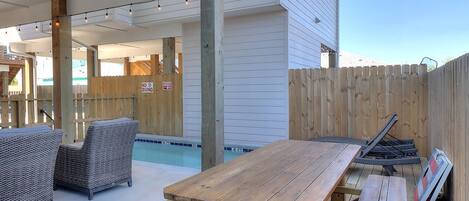 An outdoor patio area with a wooden table and chairs, a pool, and a wooden fence.